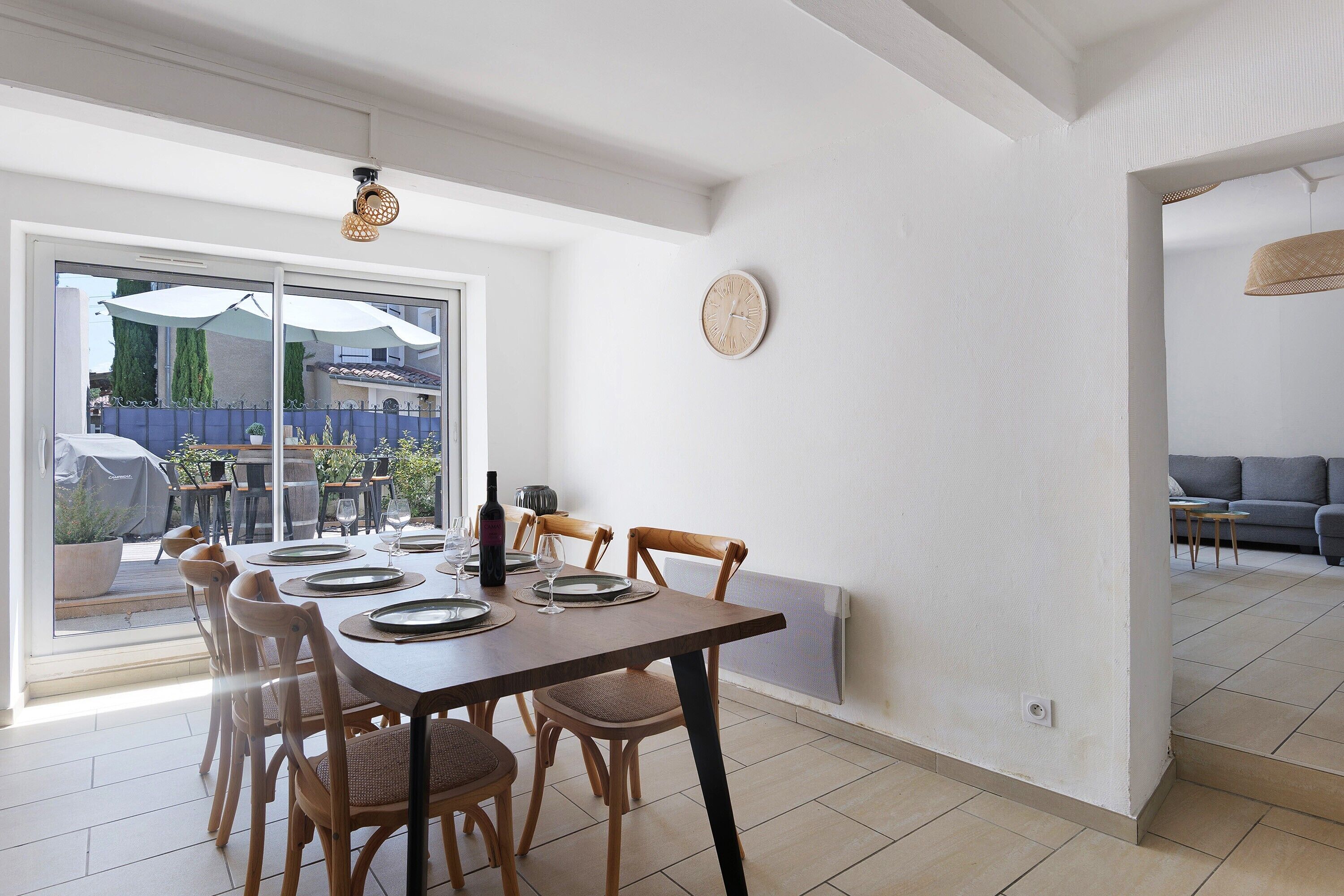 A dining area with a table set for four, a clock on the wall, and a view through a glass door to the outside.