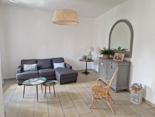 A living room with a grey sectional sofa, a small round table, a wicker chair, a cabinet with a mirror, and a hanging light fixture.
