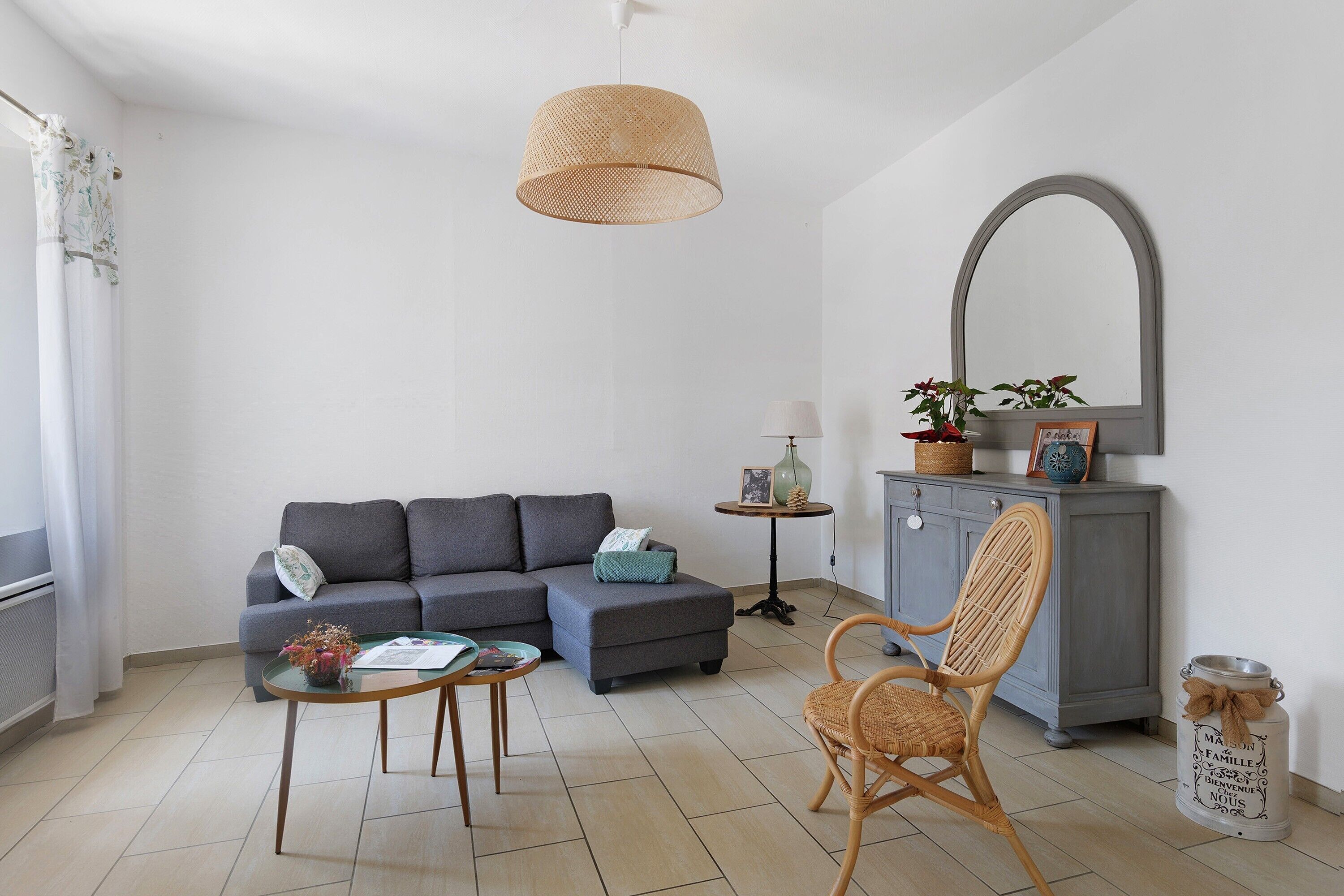 Un salon comprenant un canapé, un pouf, deux petites tables, un fauteuil, un luminaire suspendu, un miroir et un meuble.