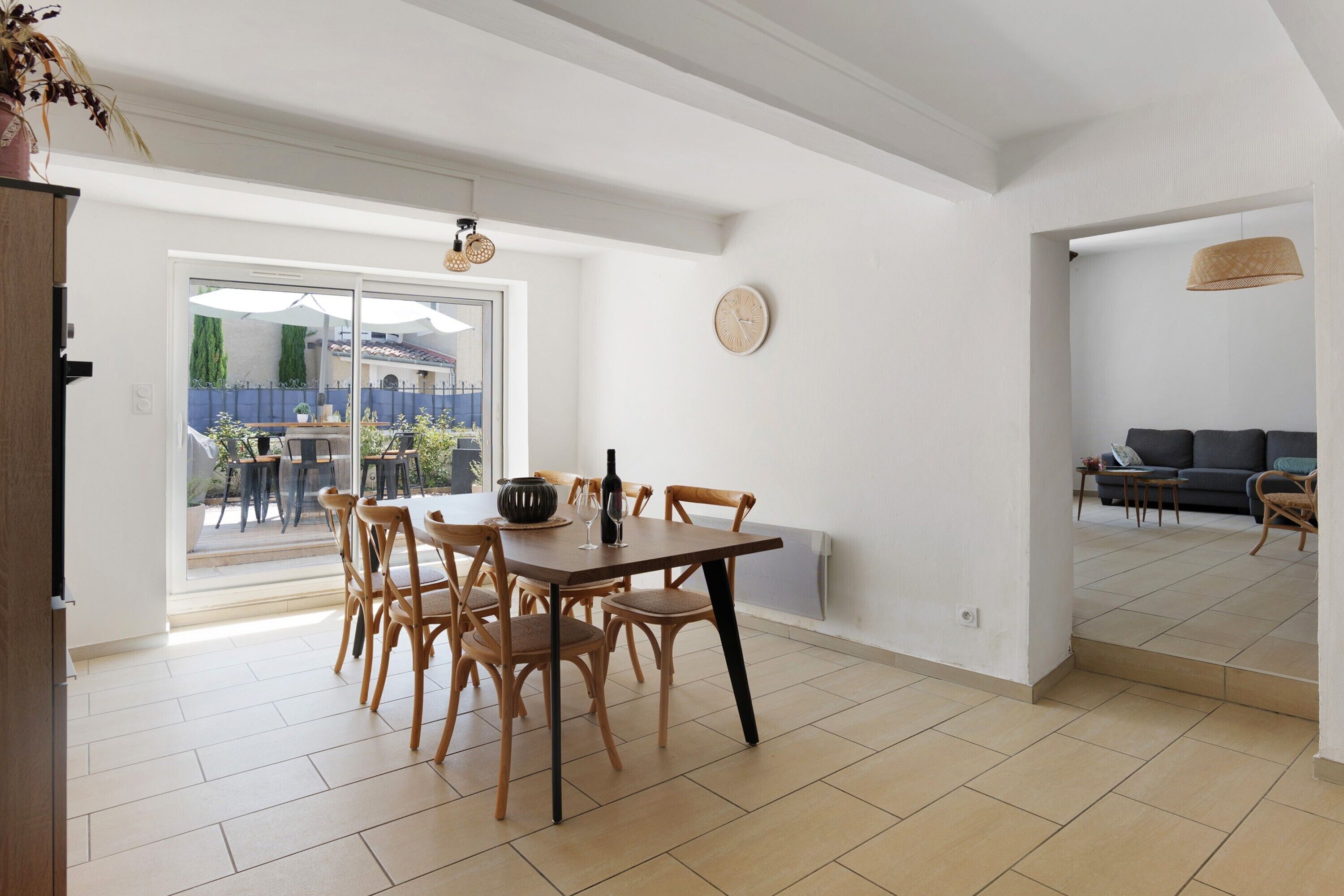 Un coin repas avec une table et des chaises, une horloge au mur et une vue sur l’extérieur à travers une porte vitrée.