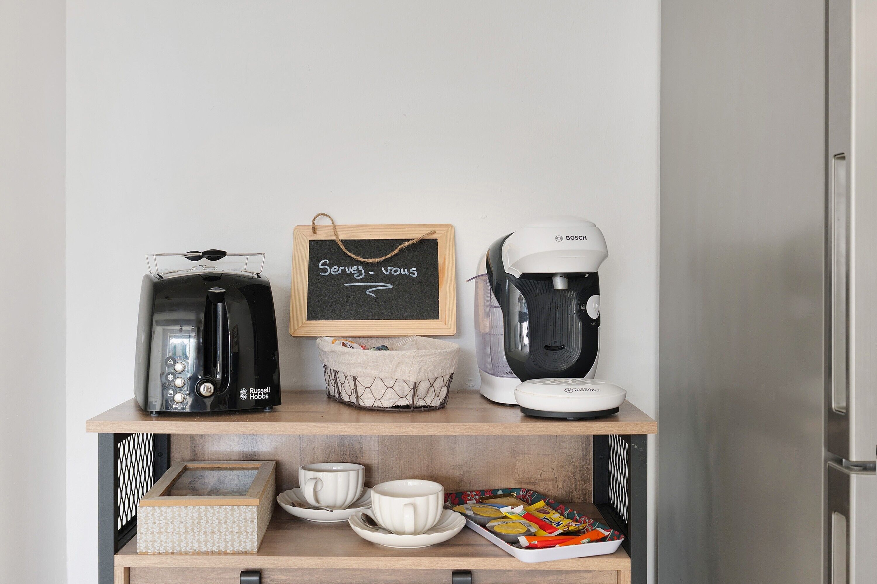 Angolo cottura con macchina del caffè, bollitore e una piccola lavagna con la scritta 'Serve - you'.