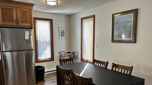 Une cuisine avec des meubles en bois, un réfrigérateur en acier inoxydable, une table à manger avec des chaises et deux fenêtres équipées de stores.