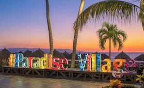 A sign with the words 'Paradise Village' in colorful letters, palm trees, and a sunset in the background. - Beautiful Condo steps from the sandy beautiful beach of Puerto Vallarta, MX. (Nuevo Vallarta)
