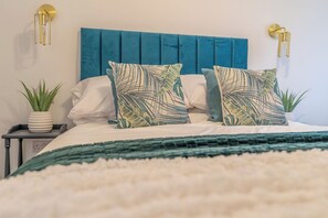 Een bed met een blauwgroen hoofdeinde, twee kussens met bladmotief, een witte deken en een groene plaid.
