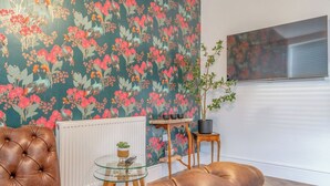 Een woonkamer met een gestofte leren bank, een glazen salontafel, een radiator, een potplant en een wand met bloemenbehang.