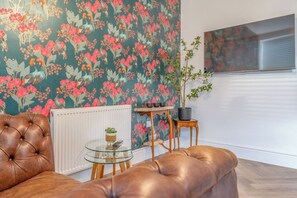 A living room with a tufted leather sofa, a glass coffee table, a radiator, a potted plant, and a wall with floral wallpaper. - Beautifully Designed 1 Bed Flat (Morecambe)