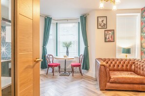 A room with a wooden door, a brown leather sofa, a small round table with two chairs, and a window with curtains. - Beautifully Designed 1 Bed Flat (Morecambe)