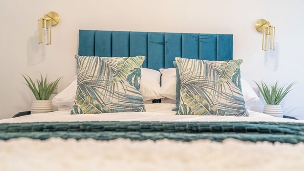 Een bed met een blauw gestoffeerd hoofdeinde, twee kussens met een bladmotief, en twee wandlampen.