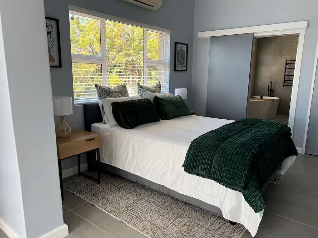 A bedroom with a bed, a bedside table, a lamp, a window with blinds, and a door leading to a bathroom.
