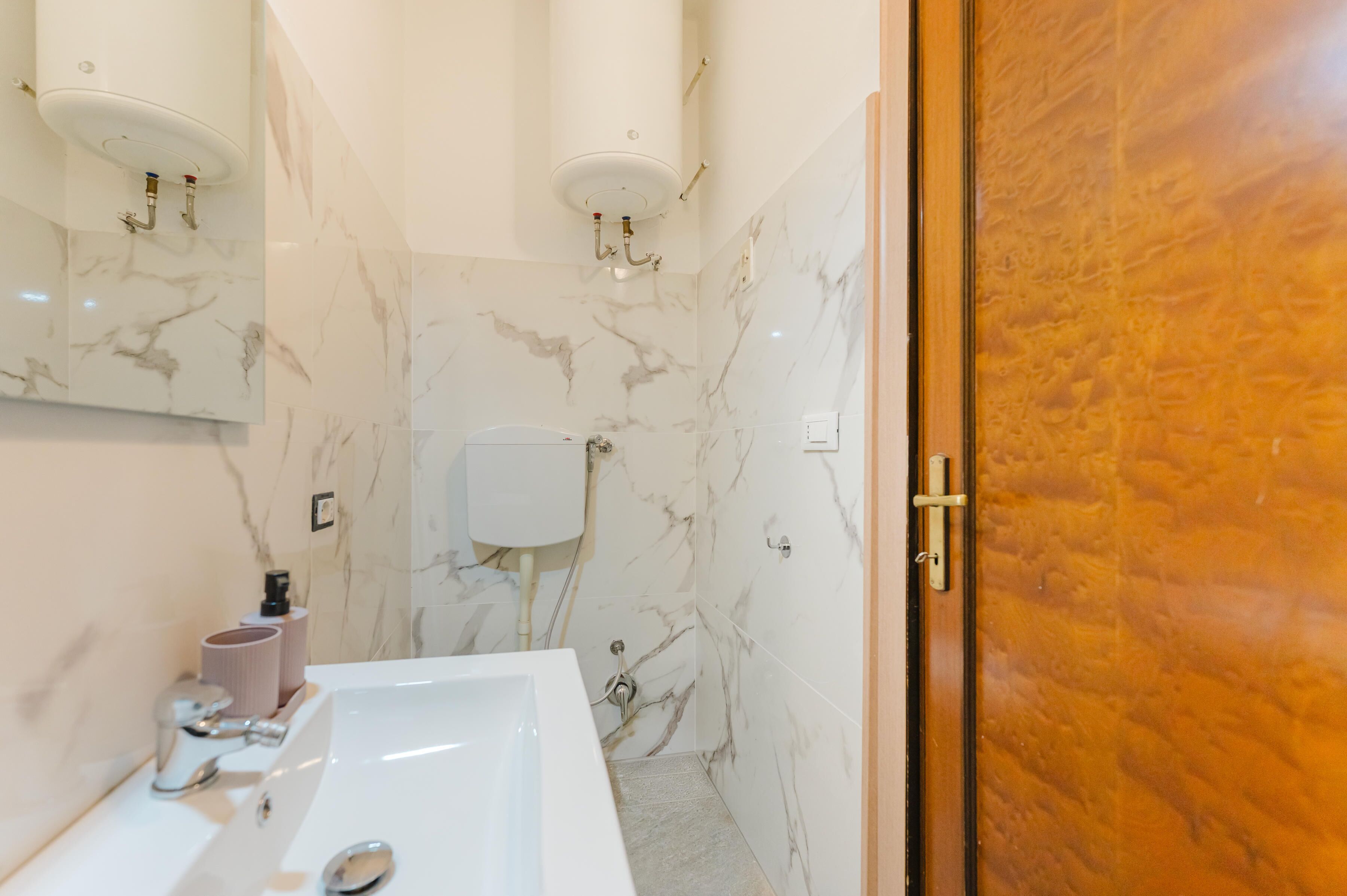 Un baño con lavamanos blanco, paredes de mármol y puerta de madera.