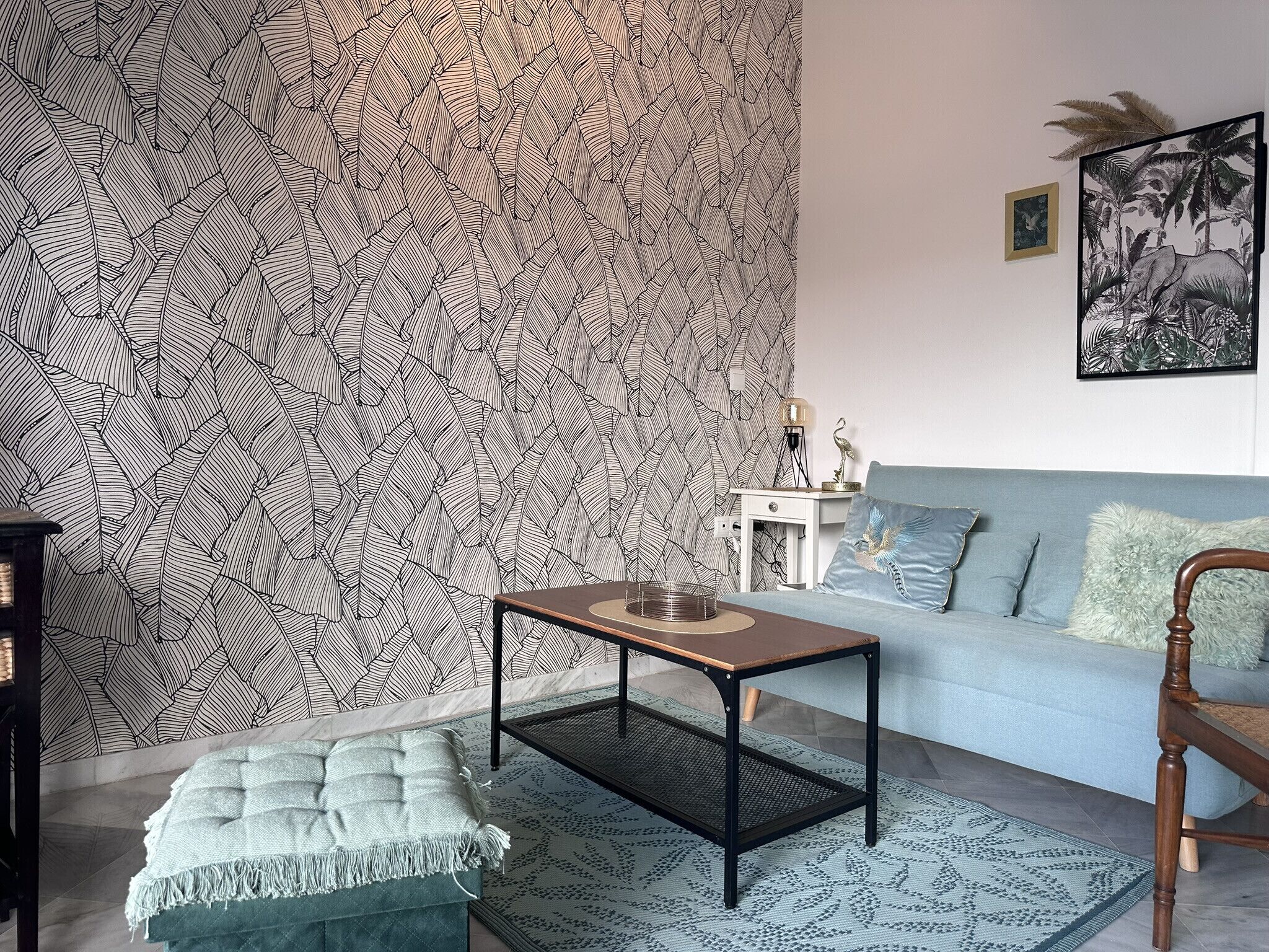 Un salon avec un papier peint à motifs, un canapé bleu clair, une table basse en bois et une petite table d’appoint blanche avec une lampe.