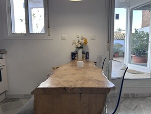 A dining area with a wooden table, chairs, a vase with flowers, and a view of the outside through large windows. - Apartamento El Convento - Ole Solutions (Jerez de la Frontera)