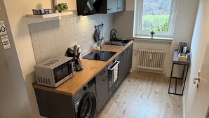 A modern kitchen with a built-in washing machine, a microwave, and a window with a view of greenery. - GNA02.01 - Apartment with 5 beds, modern kitchen, balcony & parking space (Lüdenscheid)