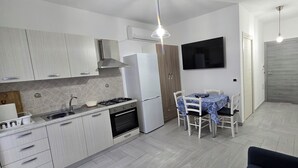 Une cuisine moderne avec des armoires blanches, une cuisinière à gaz et un four encastré. On y trouve un coin repas avec une table et des chaises, ainsi qu’un téléviseur fixé au mur.