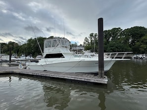 A white boat docked at a marina with a wooden dock, surrounded by calm water and trees.