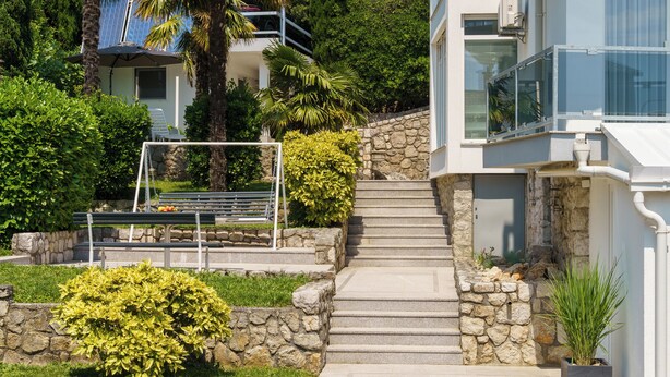 Un edificio moderno con una scalinata in pietra che conduce a un balcone, immerso nel verde sotto un cielo sereno.