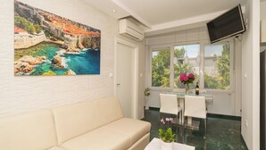 Una stanza con un divano color beige, un tavolo da pranzo con sedie, una TV montata a parete e una finestra vista sul verde.