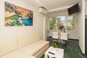 Ein Zimmer mit einem beigen Sofa, einem Esstisch mit Stühlen, einem an der Wand montierten Fernseher und einem Fenster mit Blick auf Grünflächen.