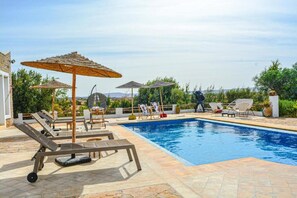 Piscina com espreguiçadeiras e guarda-sóis, cercada por vegetação e céu azul.