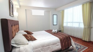 Una habitación de hotel con una cama grande, cabecero, ventana con cortinas y una lámpara fijada a la pared.