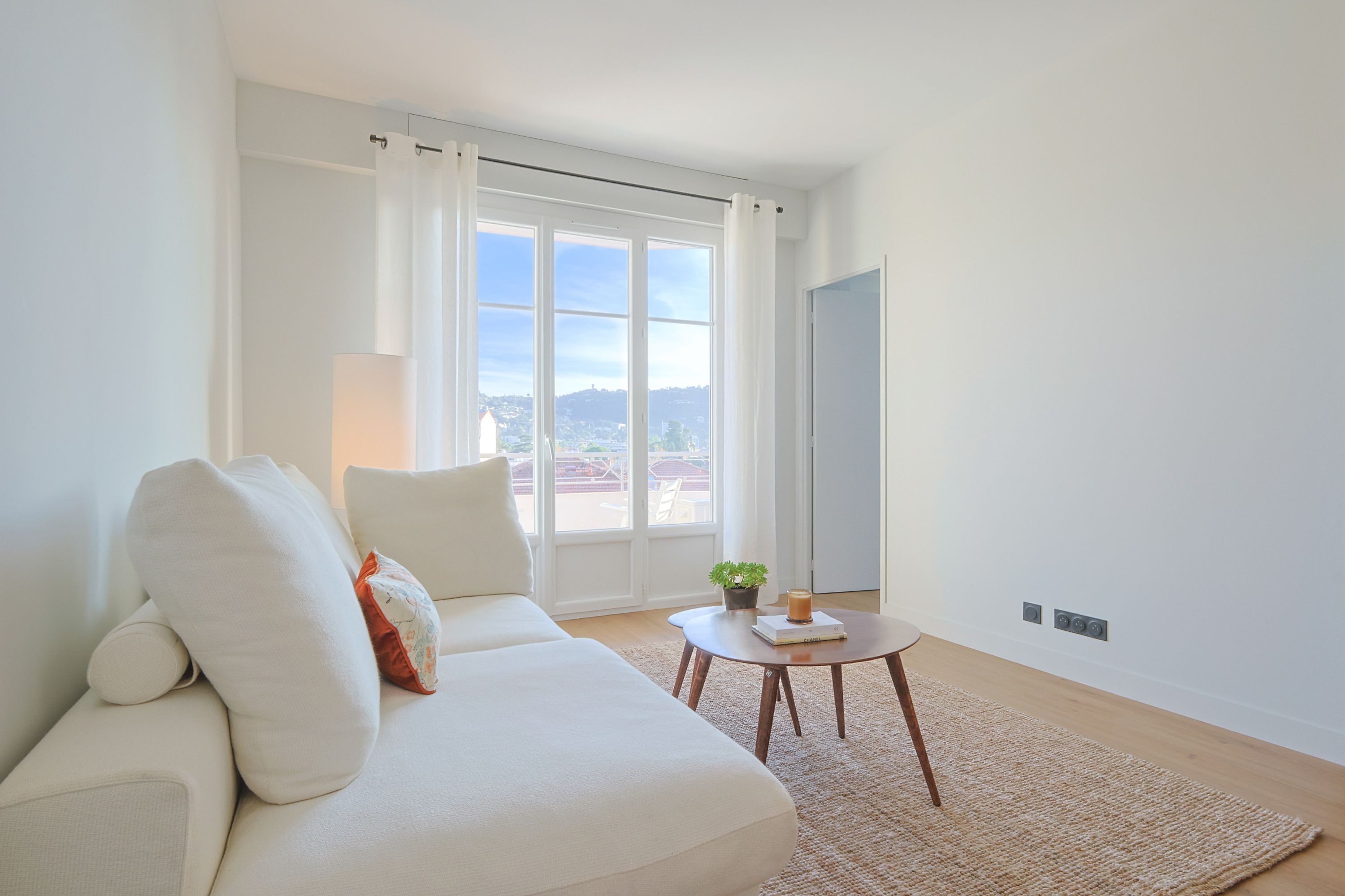 Uma sala de estar moderna com um sofá branco, uma mesinha redonda com um livro e uma planta, e vista através de grandes janelas.