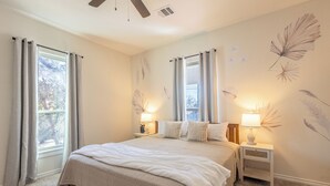 Un dormitorio con una cama grande, dos mesitas de noche con lámparas, un ventilador de techo y una ventana con cortinas.
