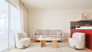 Ein modernes Wohnzimmer mit einer Couch, zwei Sesseln, einem Couchtisch, einer Küchenzeile mit roter Akzentwand und einem großen Fenster.