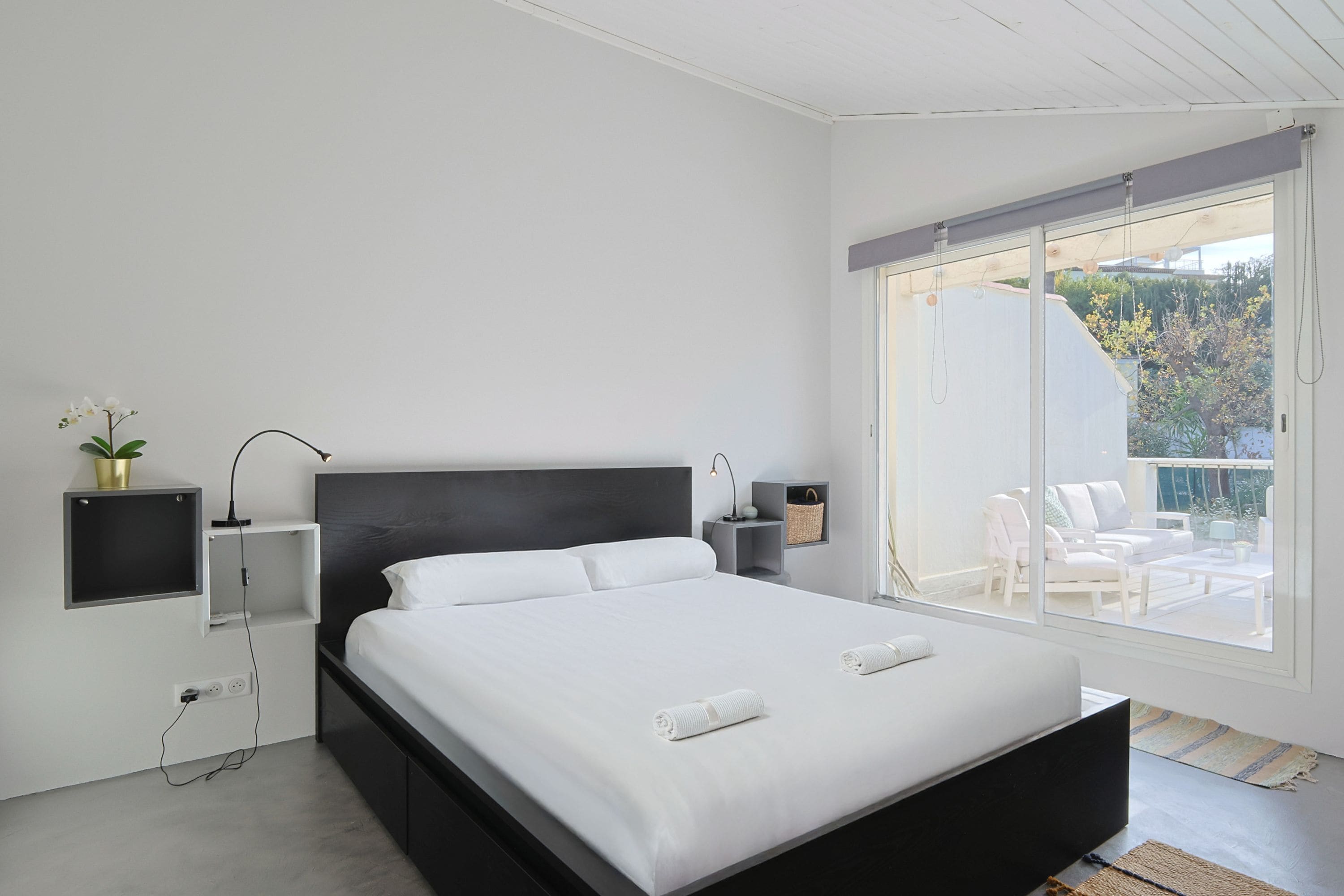 A modern bedroom with a large bed, bedside tables, a wall-mounted shelf, and a sliding glass door leading to an outdoor seating area.