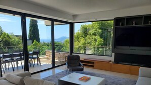 A modern living room with a large TV, comfortable seating, and a view of greenery and mountains. - 004 Villa Le Palm in Le Cannet (Le Cannet)