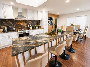 A modern kitchen with a marble countertop, white cabinets, and a dining area with bar stools. - Luxury 4BR Family Retreat w/Pool Sleeps 10 6 Beds (Dallas)