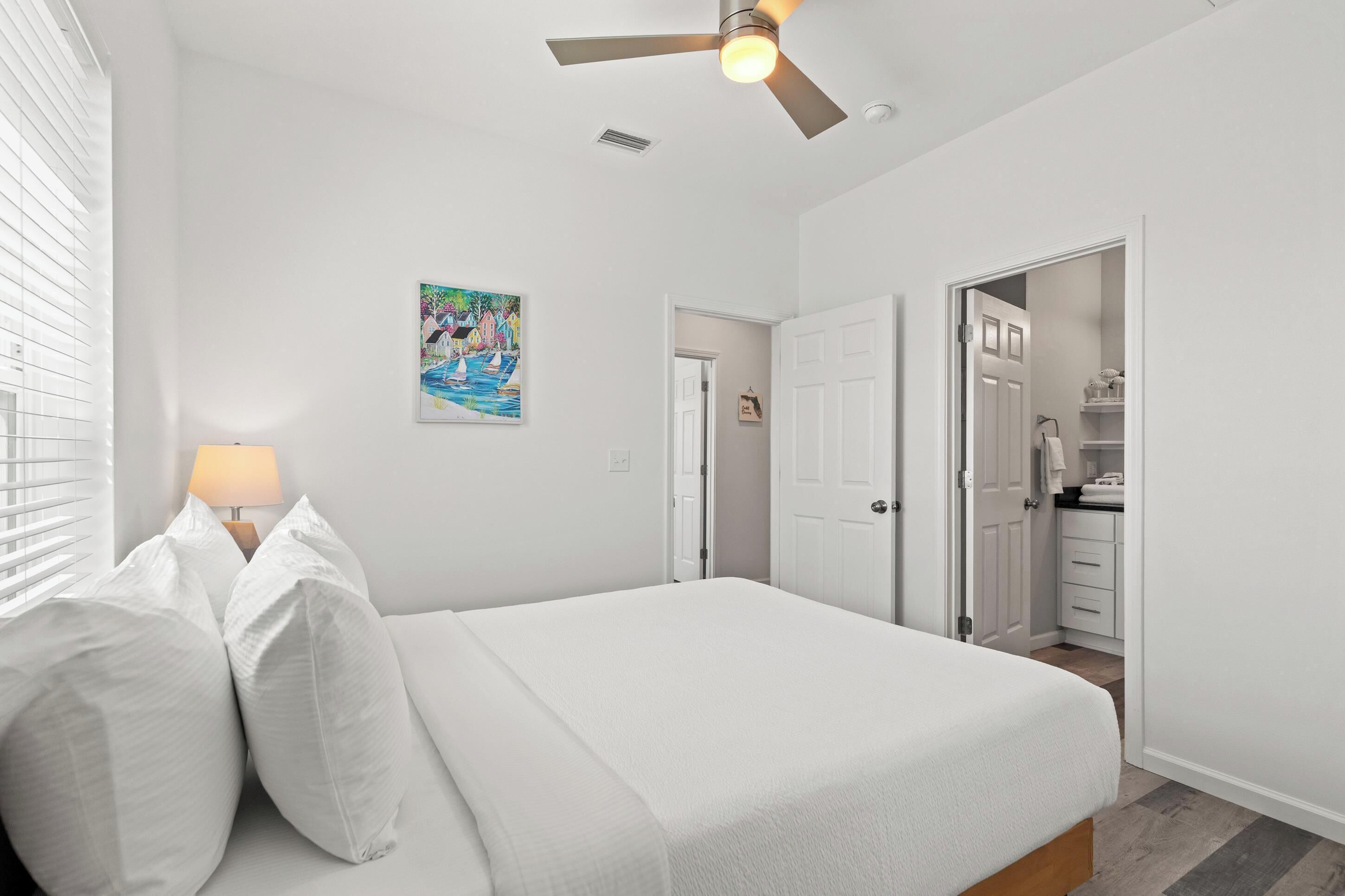 Ein Schlafzimmer mit einem Bett, einem Deckenventilator, einem Gemälde und Blick ins Badezimmer.