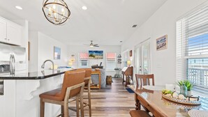 A spacious kitchen with a central island, wooden chairs, a dining table with a tray of fruits, and a view of the outdoors through large windows. - Beach House - Coastal Dreaming (Port St. Joe)