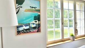 Ein Zimmer mit großem Fenster, einem gerahmten Kunstwerk an der Wand, einer Lampe und einer Pflanze auf der Fensterbank.