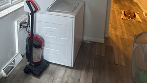 A compact laundry room with a stacked washer and dryer, a vacuum cleaner, and a window with a metal grille. - Tranquil 3-bedroom apartment with AC in Pittsburgh Oakland area. (Pittsburgh)