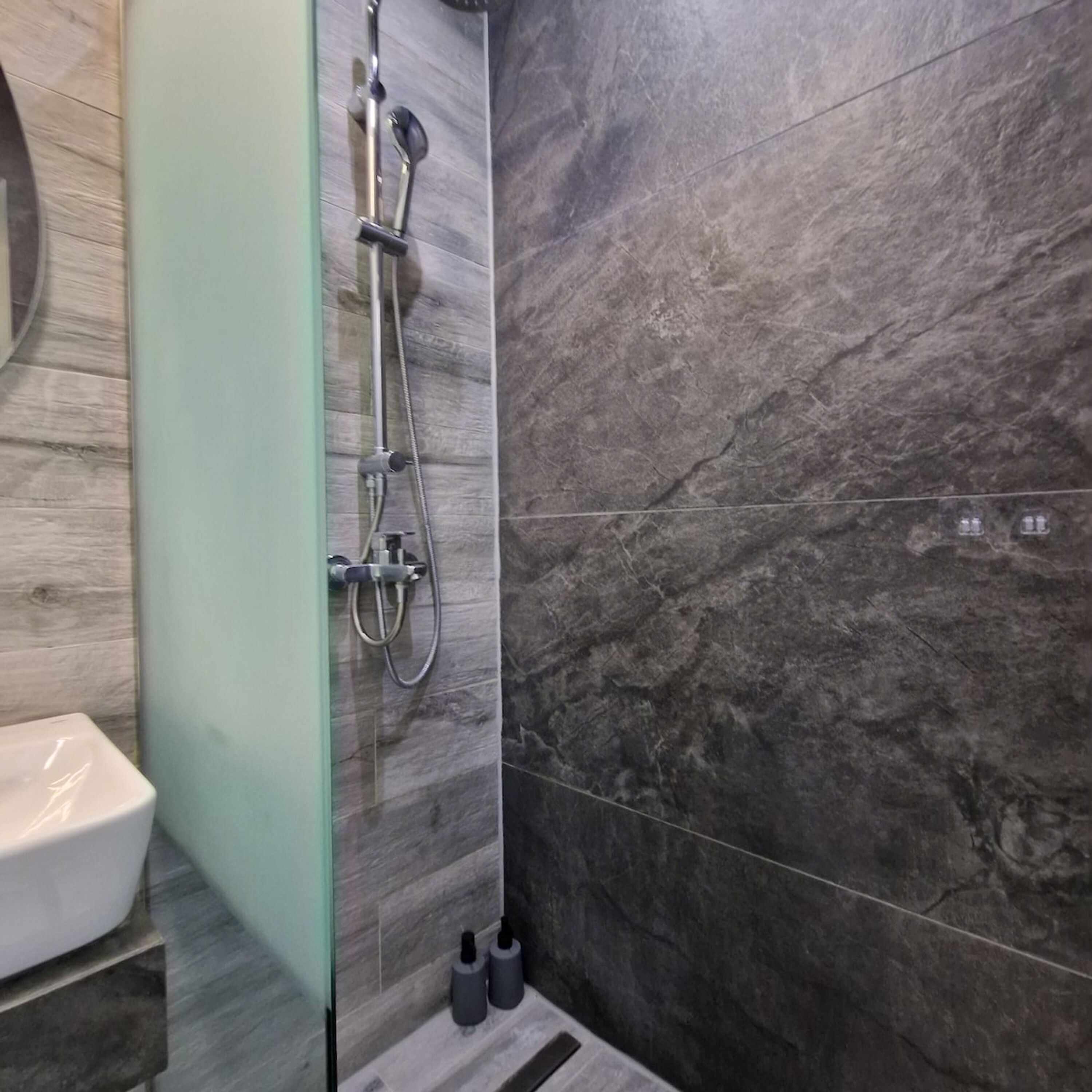 A modern bathroom with a glass shower enclosure, a white sink, and dark marble tiles.