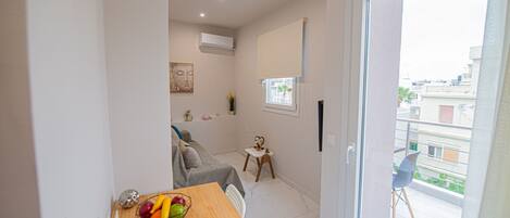 A compact living space with a dining area, a sofa, a small table with a fruit bowl, a window, and a balcony with a chair.