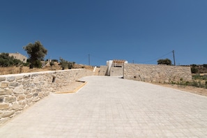 Ein gepflasterter Weg führt zu einem Steinbau mit moderner Dachkonstruktion, umgeben von trockener Vegetation und blauem Himmel.