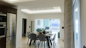 Una cocina moderna con horno integrado y refrigerador, un comedor con mesa y sillas, y vista a una sala de estar a través de un vano abierto.
