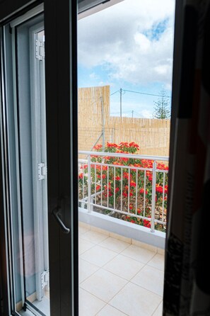 Eine offene Schiebetür führt zu einem Balkon mit Blick auf einen Holzzaun und rote Blüten.