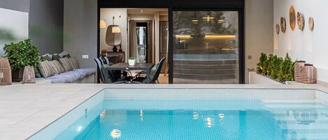 Et moderne poolområde udendørs med spisebord og stole, en sofa samt en skydedør til et køkken.