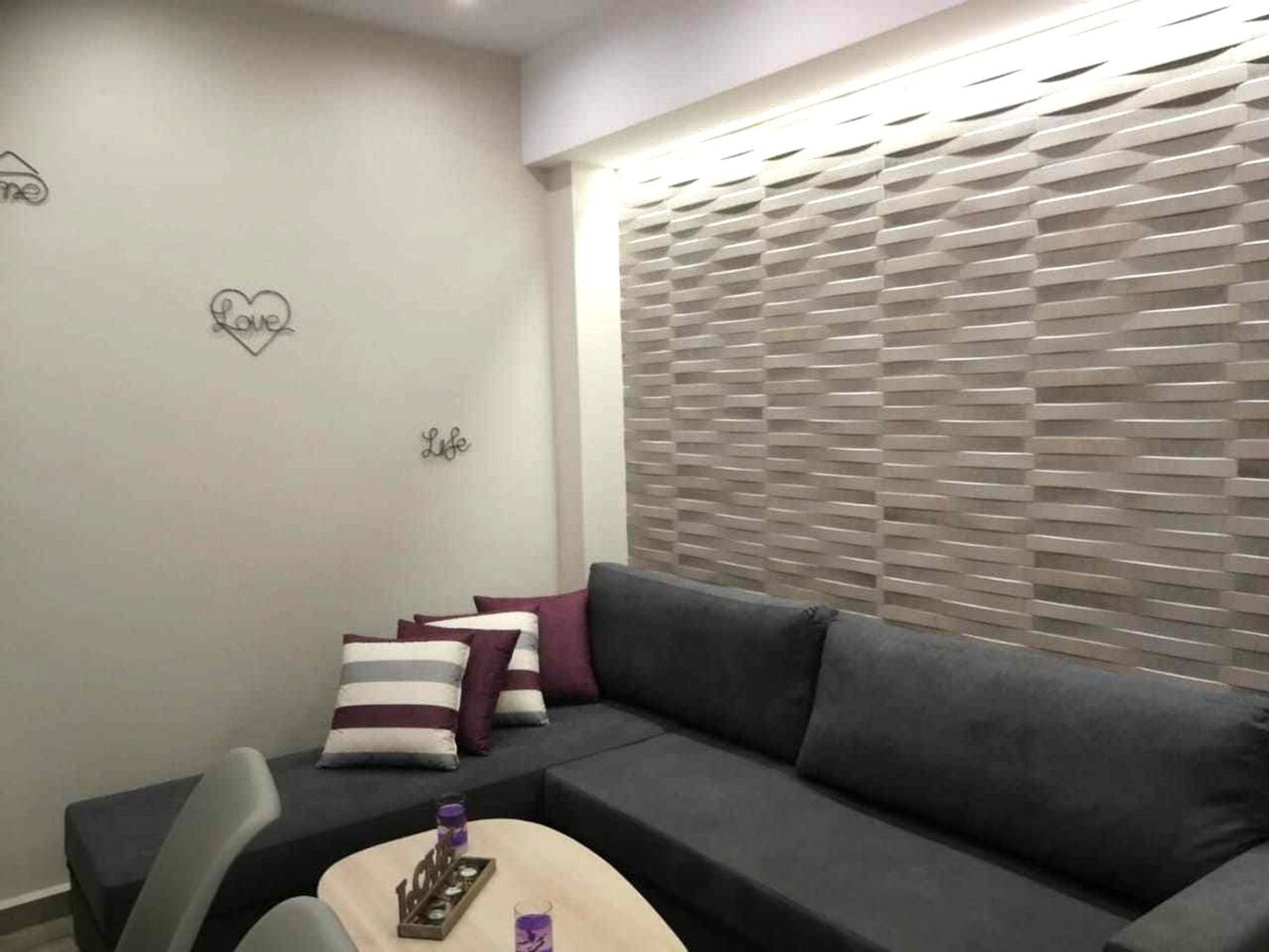 Ein modernes Wohnzimmer mit einem grauen Sofa, gestreiften Kissen, einem kleinen Beistelltisch und einer Wand mit herzförmiger Dekoration.