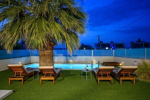 Un área junto a la piscina con sillones de descanso, una mesita con una botella y una palmera.