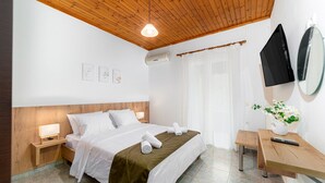 Ein Hotelzimmer mit Holzboden, einem Bett mit weißen Leinen, einem Nachttisch mit Lampe, einer kleinen Holzbank und einem an der Wand befestigten Fernseher.