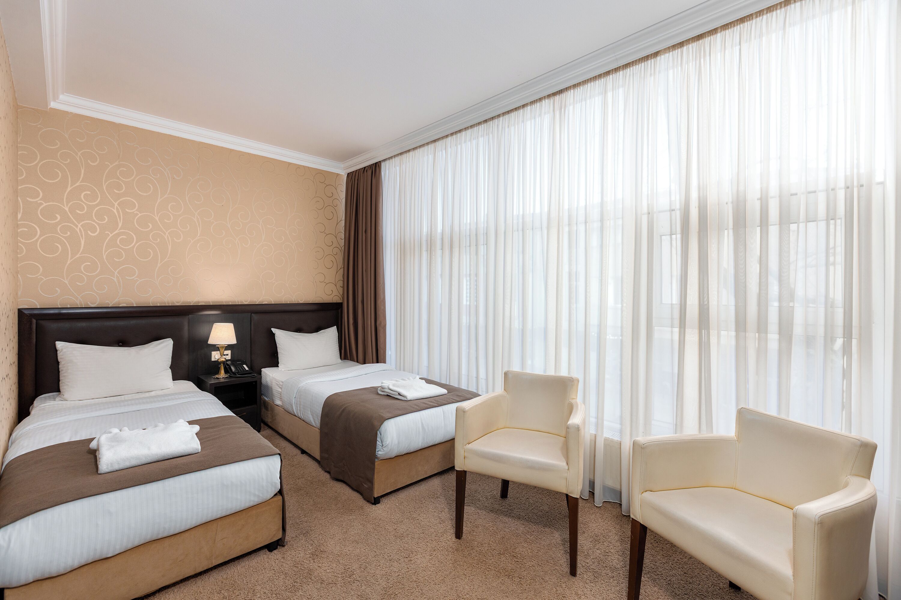 A hotel room with two beds, a chair, a nightstand, and a window with curtains.