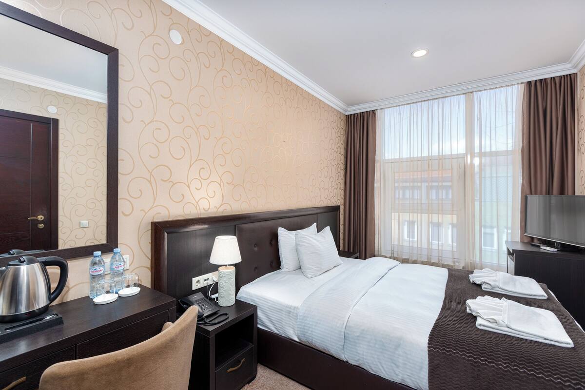 A hotel room with a bed, a desk with a telephone, a television, and a window with curtains.