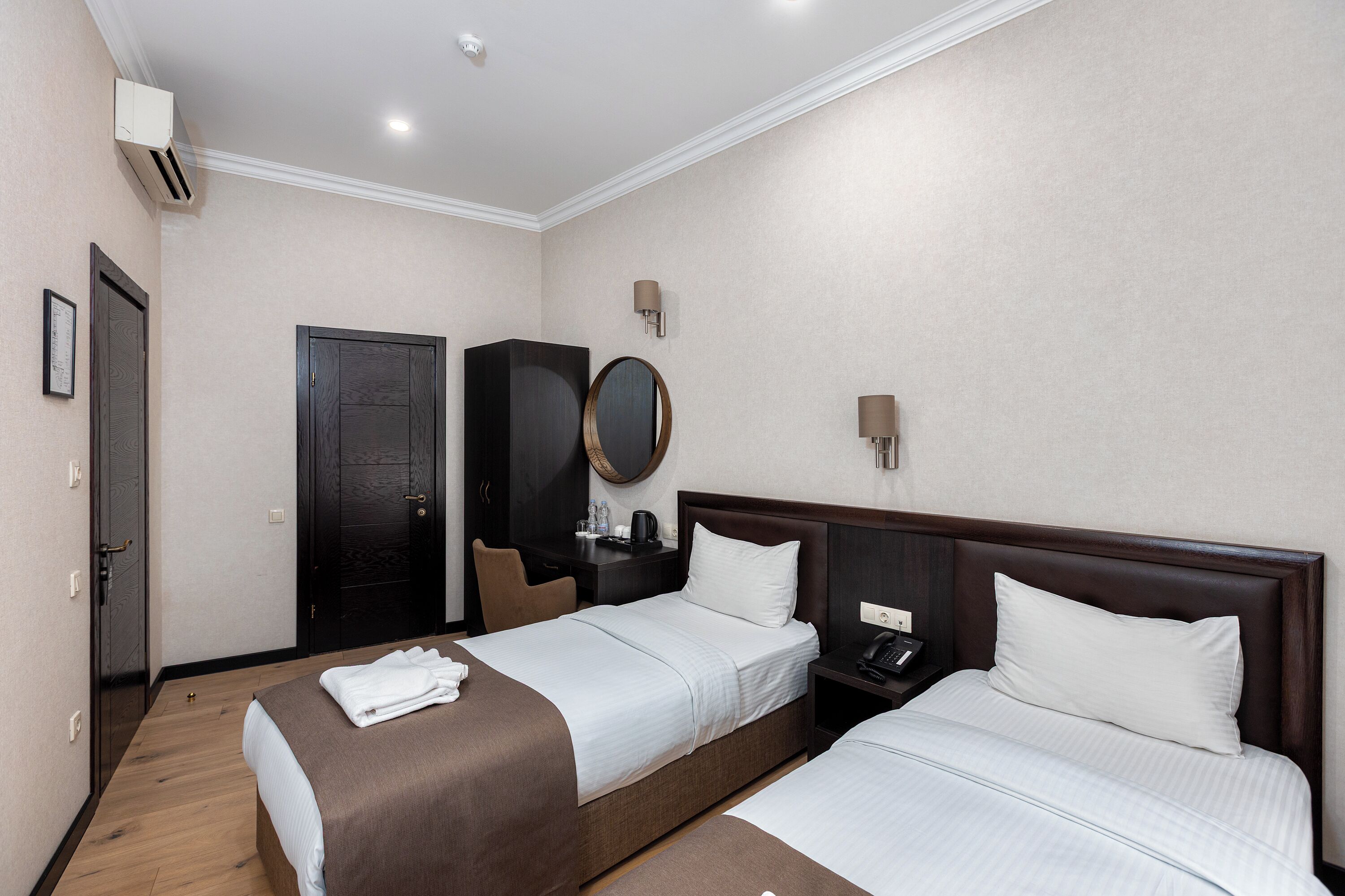 A hotel room with two beds, a desk, a wardrobe, a mirror, and a wall-mounted phone.