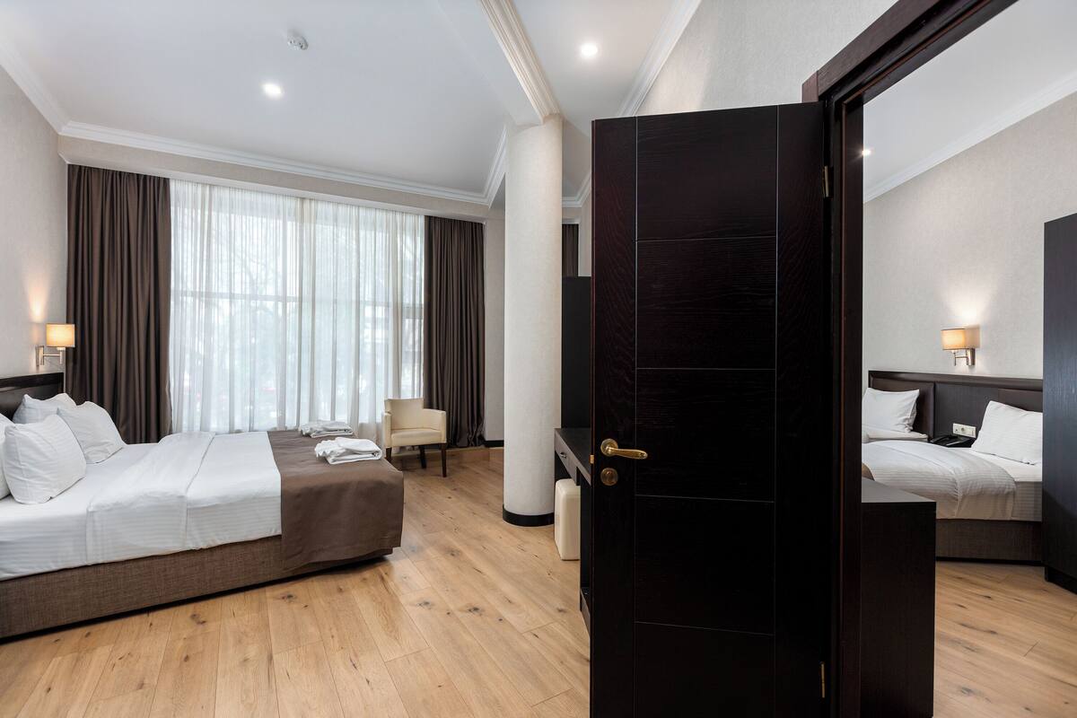 A hotel room with two beds, a sitting area, a large window with curtains, and a dark wooden door.