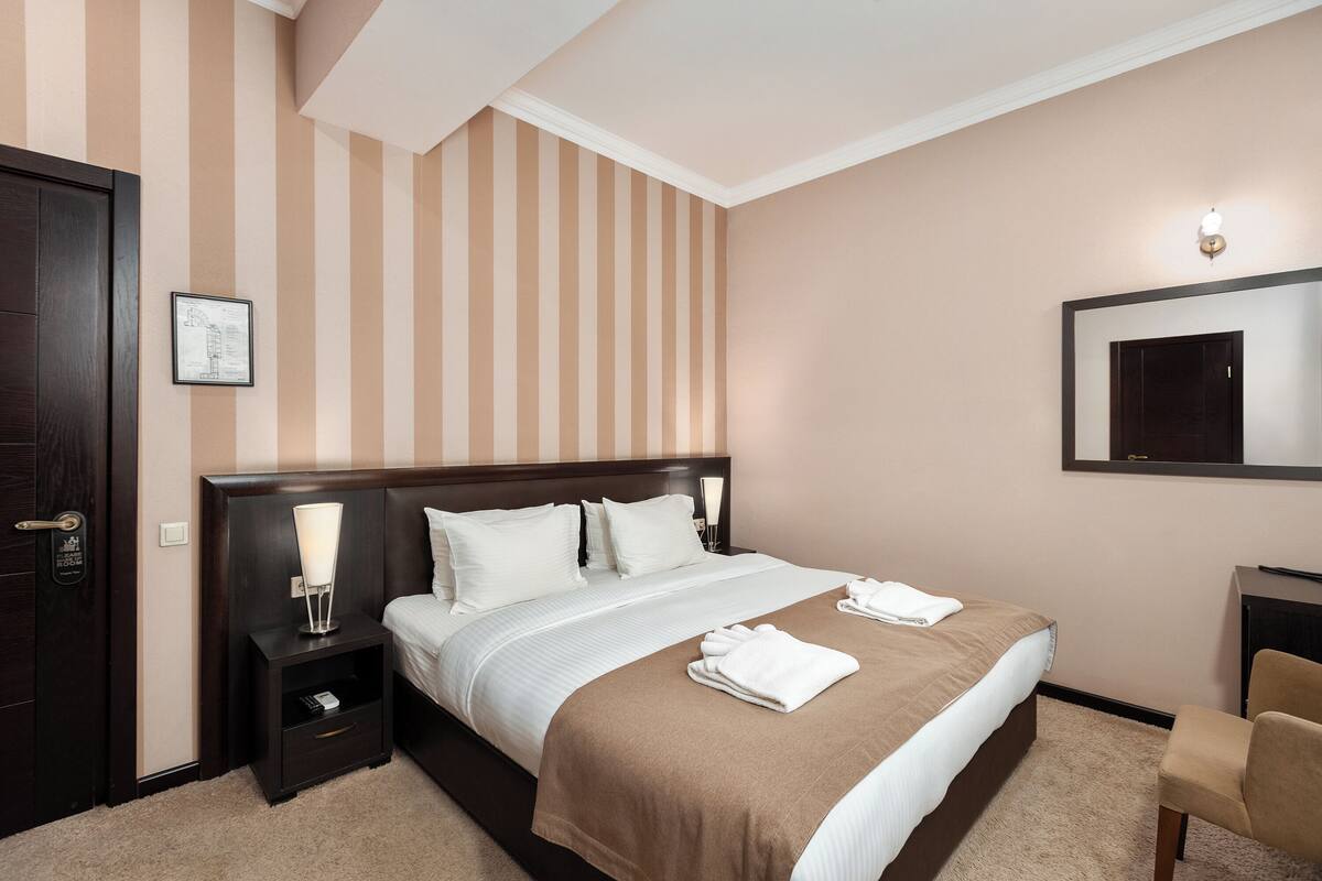 A hotel room with a bed, a nightstand, a chair, a desk, a mirror, and a door.