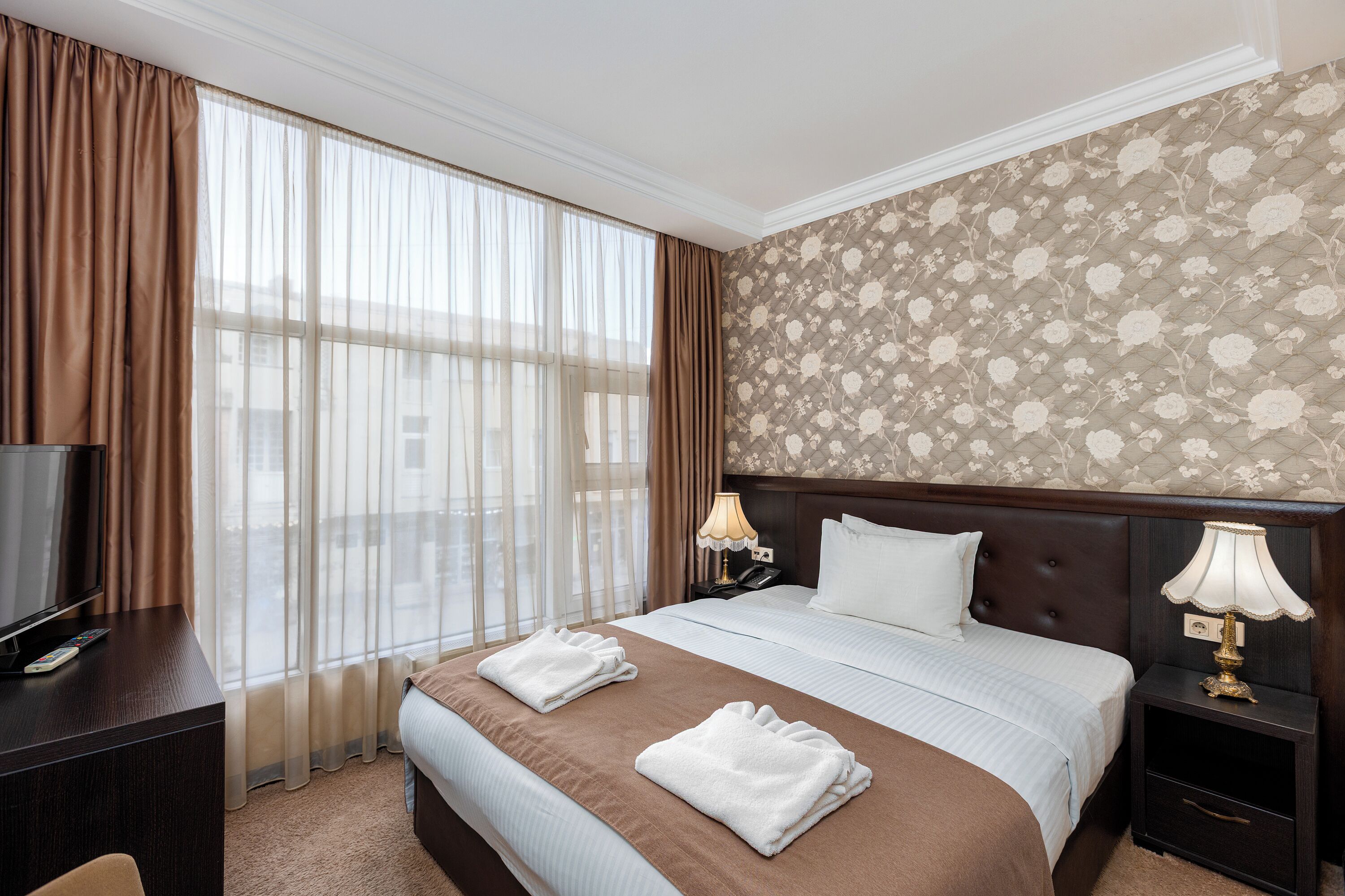 A hotel room with a large bed, a TV on a desk, a nightstand with a lamp, and a window with curtains.