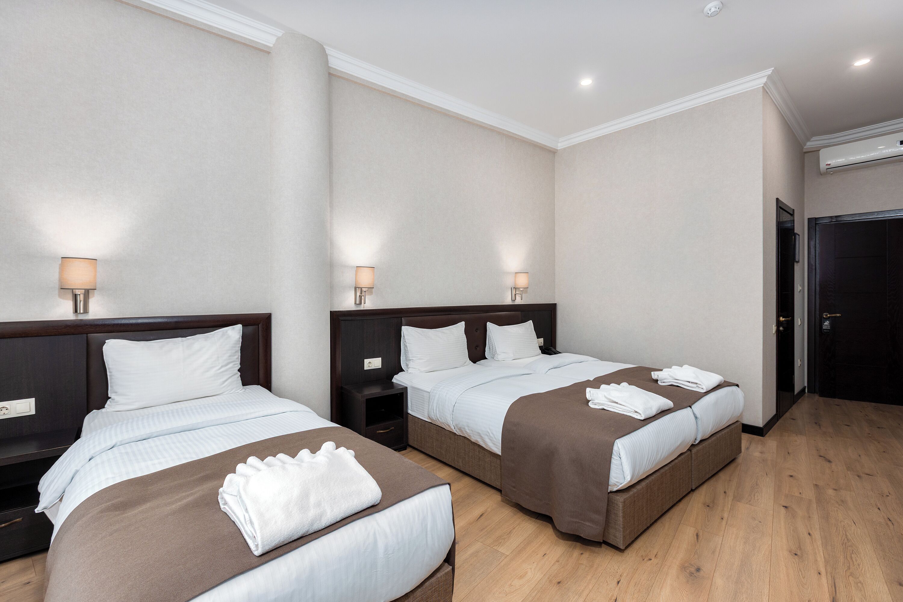 A hotel room with two beds, a wooden floor, a dark door, and a white wall.